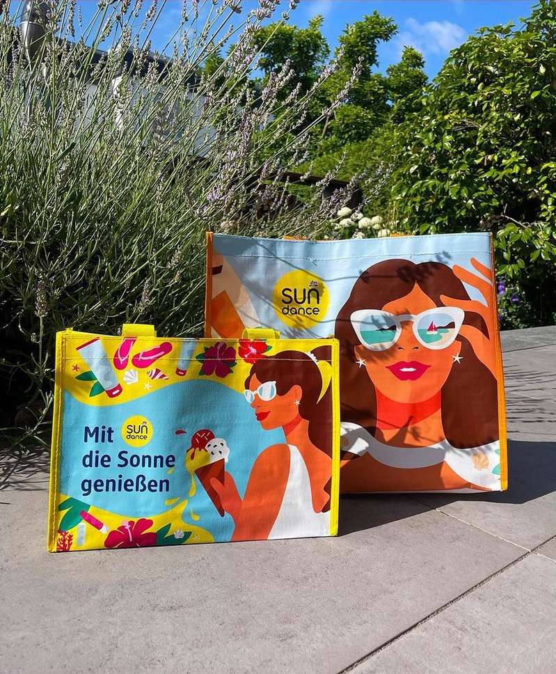 Two vibrant tote bags featuring illustrations of women in sunglasses and beach scenes sit outdoors against a backdrop of green plants.