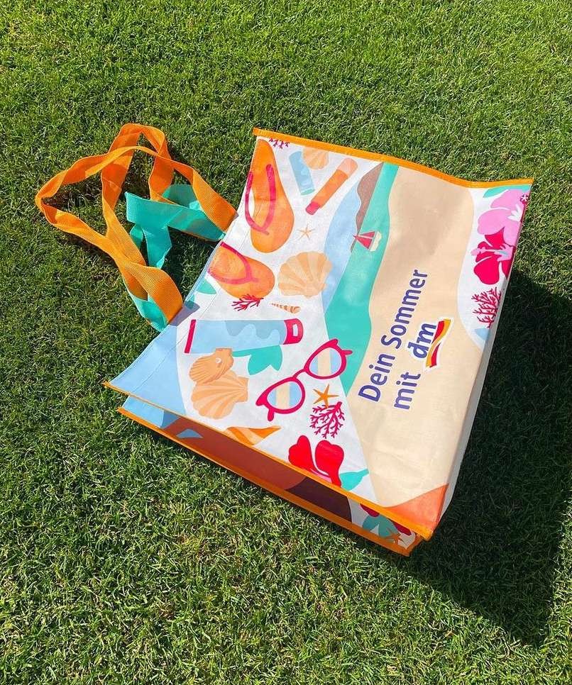 Summer-themed tote bag featuring beach-related graphics on green grass outdoors, bright sunlight, and orange and teal straps.