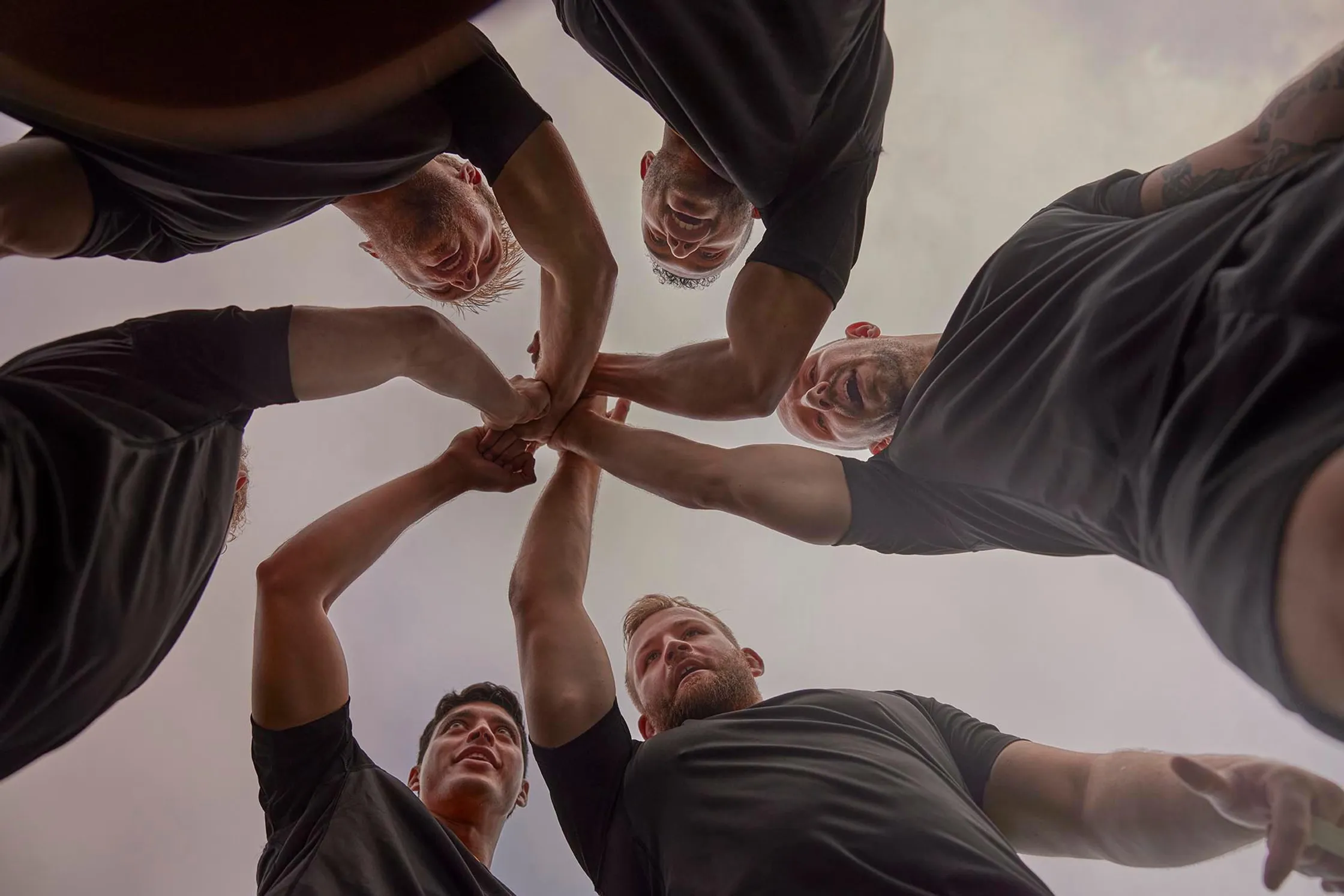 Group of six athletic men in black t-shirts huddle with arms raised, shot from a low angle against a diffused, pale sky, team spirit.