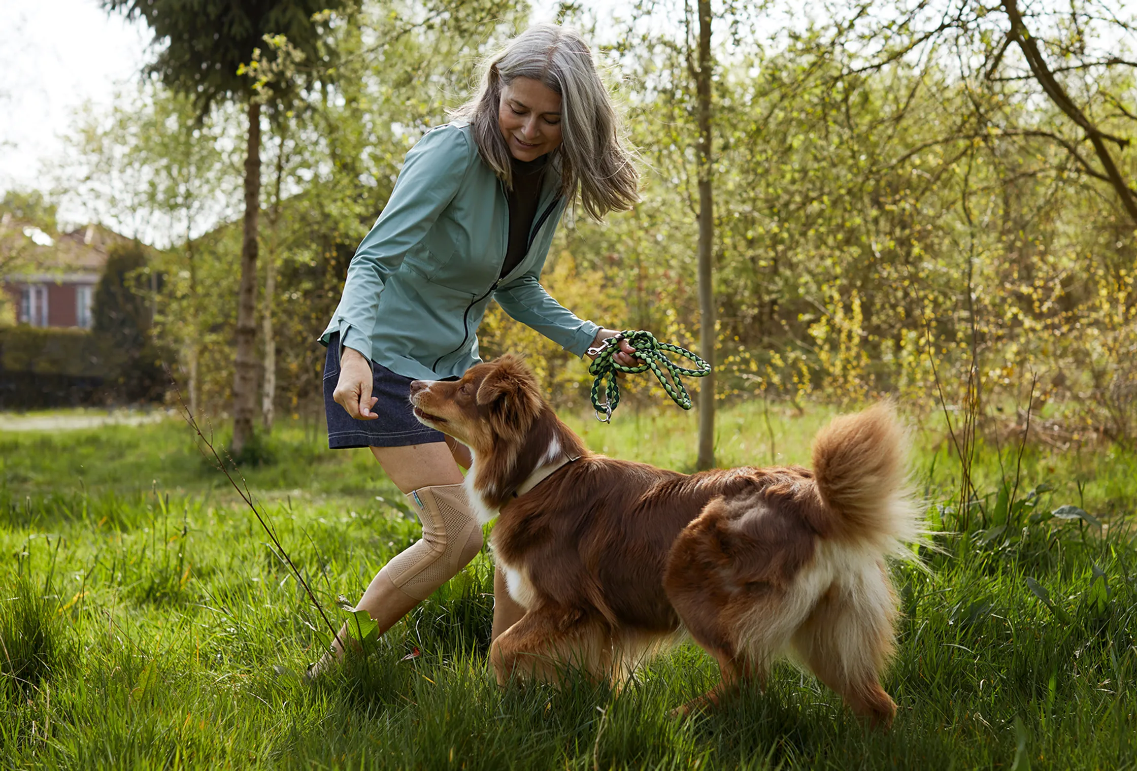 A woman plays with her dog in a grassy park, bathed in sunlight. She holds a leash, creating a lively scene of human-animal interaction outdoors.