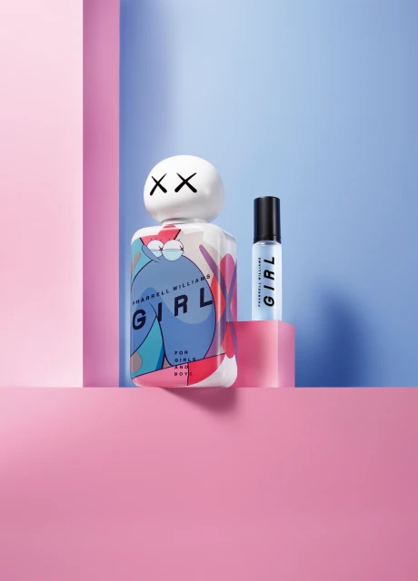 Two Pharrell Williams' Girl fragrance bottles, one with a KAWS-designed cap, displayed on pink and blue geometric platforms in a studio setting.