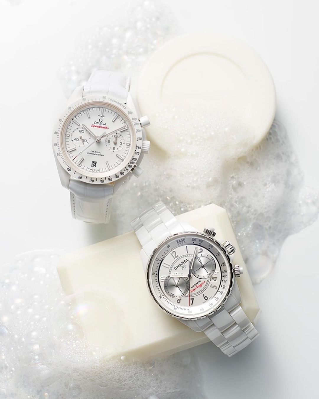 Two luxury watches from Omega and Chanel are showcased in a bright studio setting, complemented by white soap and scattered soap bubbles.