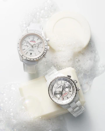 Two luxury watches from Omega and Chanel are showcased in a bright studio setting, complemented by white soap and scattered soap bubbles.