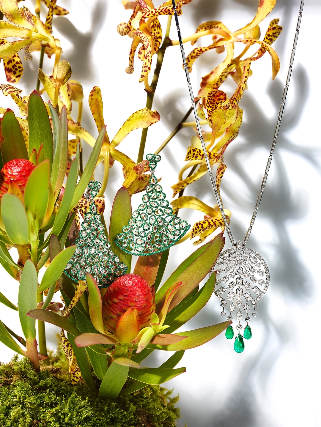 Elegant jewelry display: a diamond necklace with emerald drops alongside diamond earrings, arranged with tropical orchids and greenery on white.