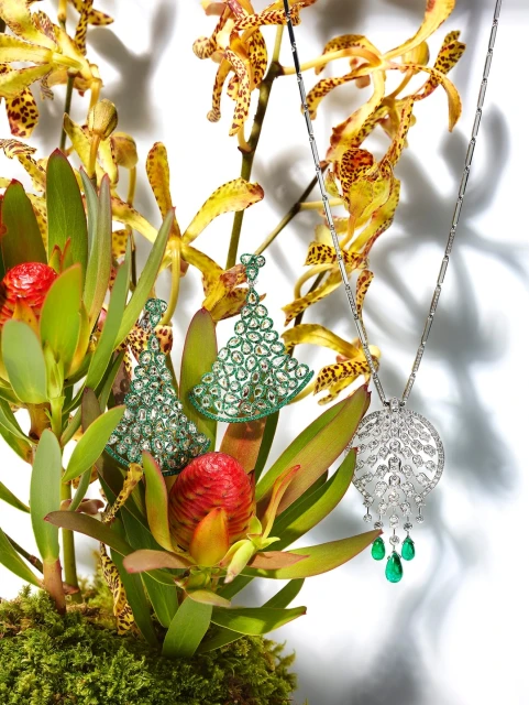 Elegant jewelry display: a diamond necklace with emerald drops alongside diamond earrings, arranged with tropical orchids and greenery on white.