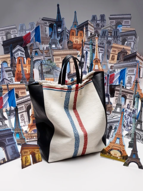 Fashion still life of a textile tote bag with black leather trim, against a backdrop of Eiffel Tower and Arc de Triomphe illustrations.