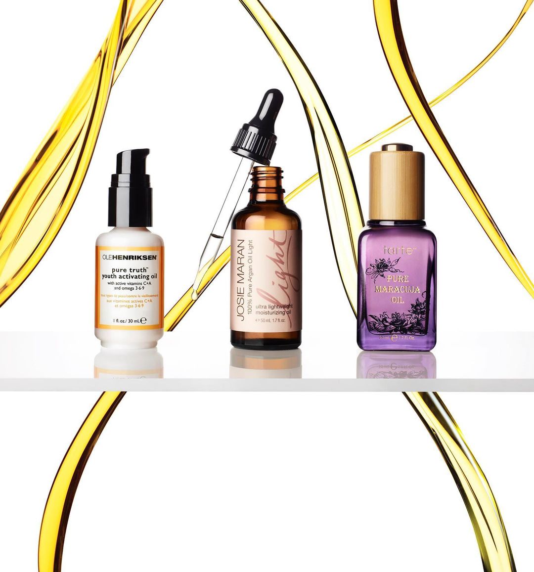 Three bottles of skincare oil from Olehenriksen, Josie Maran, and PURE Maralina stand on a shelf against a white backdrop with golden oil streams.