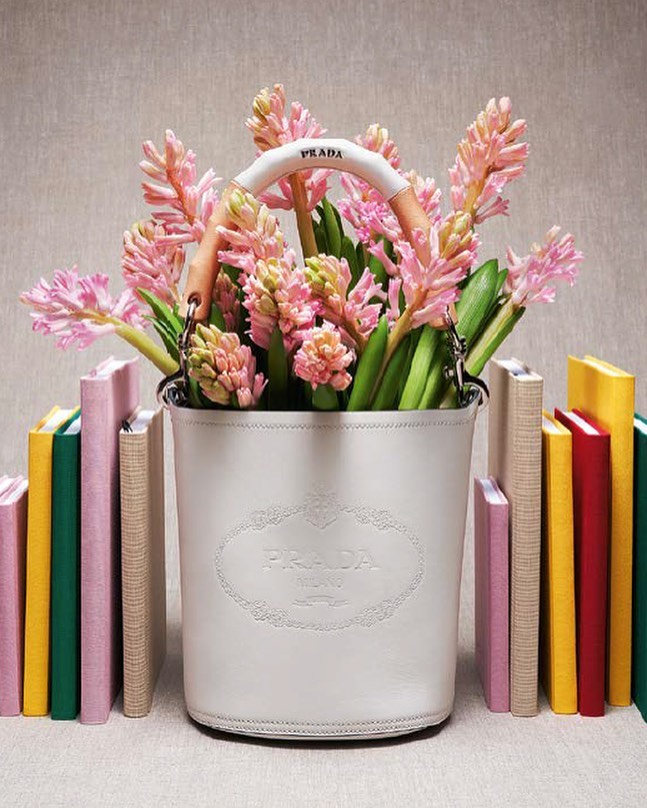 Floral arrangement featuring pink hyacinths in a white Prada bucket bag, artfully positioned between rows of colorful books.