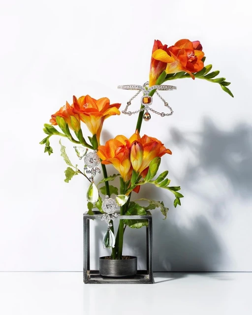Floral arrangement with orange freesia in a dark metal frame, adorned with sparkling silver jewelry on a white background.