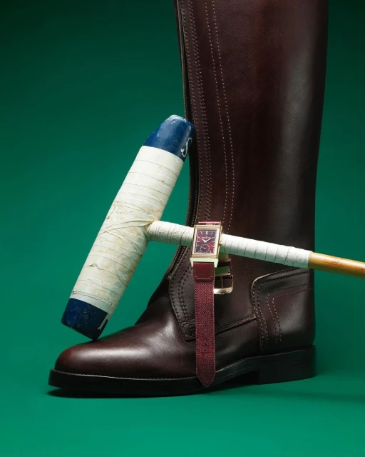Still life photo featuring a riding boot, a polo mallet, and a gold wristwatch against a solid green backdrop, product photography.