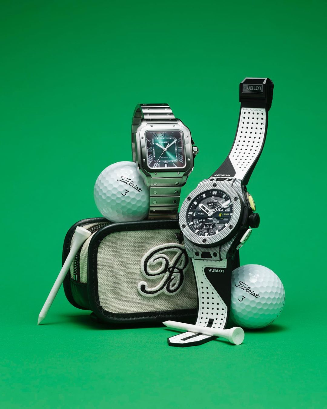 Two luxury watches sit with golf balls and golf tees on a canvas golf bag, all presented on a green surface and against a green backdrop.