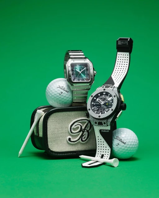 Two luxury watches sit with golf balls and golf tees on a canvas golf bag, all presented on a green surface and against a green backdrop.