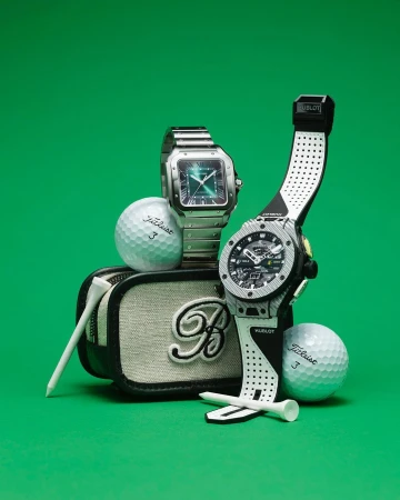 Two luxury watches sit with golf balls and golf tees on a canvas golf bag, all presented on a green surface and against a green backdrop.