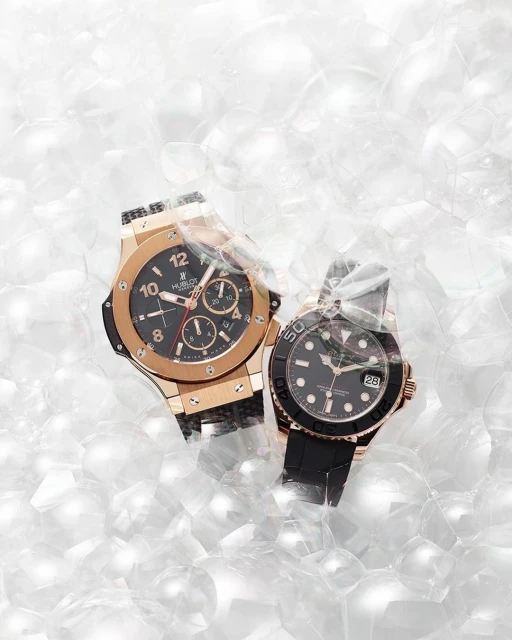 Two luxury watches, one gold and one gold with black band, are displayed amidst a backdrop of translucent white bubbles.
