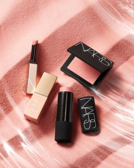 NARS makeup products including lipstick, blush, and lip color are displayed against a textured pink background with light reflections.