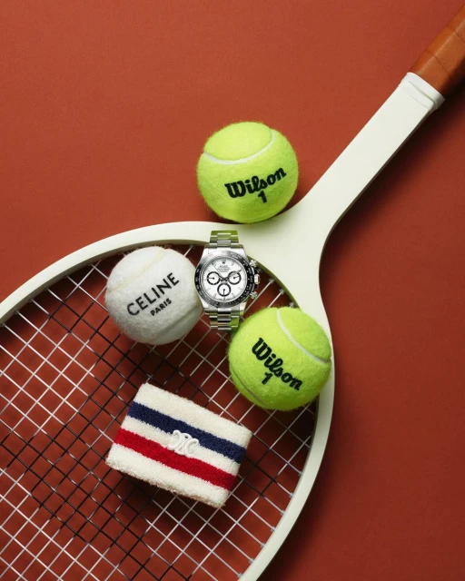 Tennis scene with a racket, balls from Wilson and Celine, a wristband, and a silver wristwatch, staged on an orange background.