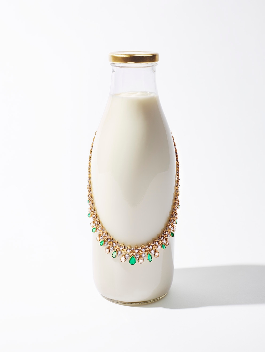A glass milk bottle on a white surface is adorned with an ornate gold, green, and white jeweled necklace in a still-life studio photograph.