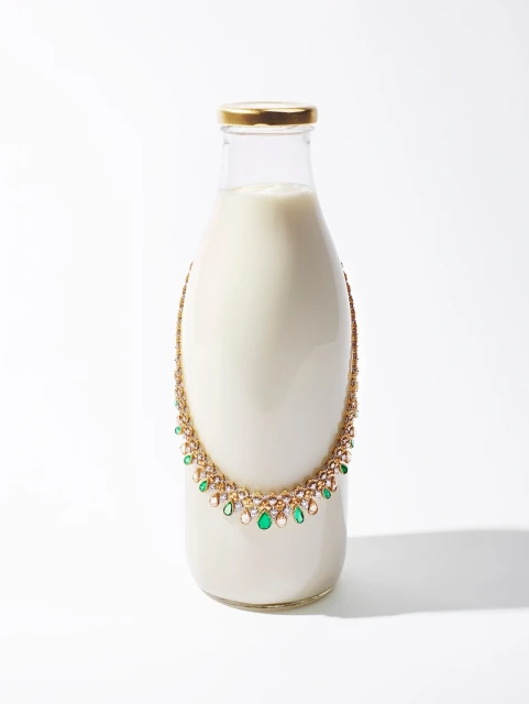 A glass milk bottle on a white surface is adorned with an ornate gold, green, and white jeweled necklace in a still-life studio photograph.