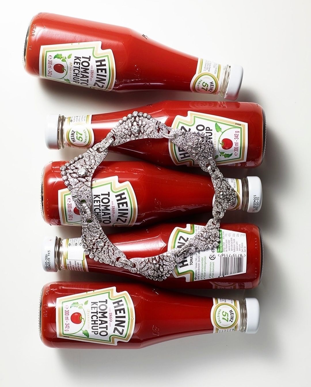 Overhead shot: Five Heinz tomato ketchup bottles, caps at alternating ends, with a diamond headband draped across the middle three.