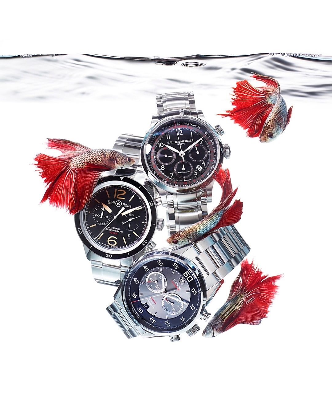 Luxury watches on metal bands are submerged in water, surrounded by red and iridescent Betta fish. A studio setup with a bright white background.