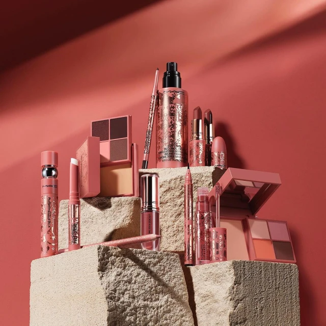A collection of MAC makeup products, including lipsticks, eyeshadow palettes, and foundation, arranged on a textured stone surface.
