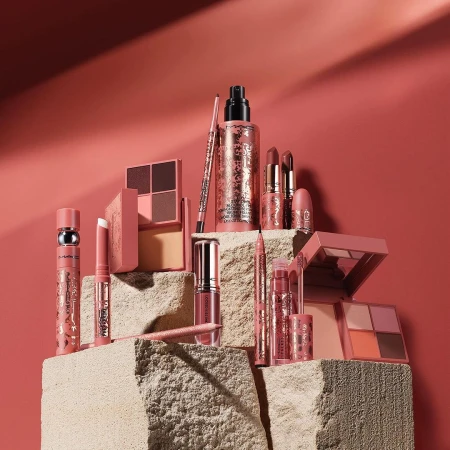 A collection of MAC makeup products, including lipsticks, eyeshadow palettes, and foundation, arranged on a textured stone surface.