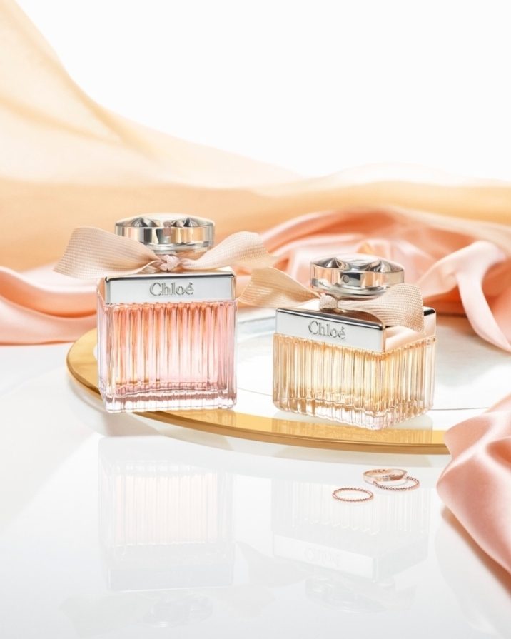 Two Chloe perfume bottles are displayed on a mirrored surface with rings, alongside draped peach satin fabric in a studio setup.