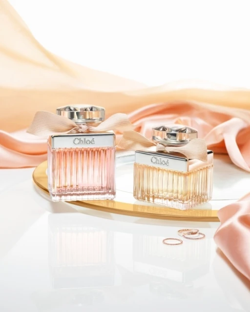 Two Chloe perfume bottles are displayed on a mirrored surface with rings, alongside draped peach satin fabric in a studio setup.