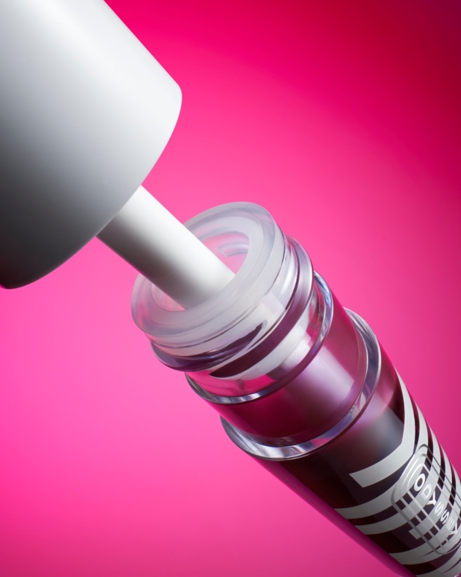 Close-up studio shot of a lip gloss bottle with a white applicator against a vibrant pink background, highlighting the product's design.