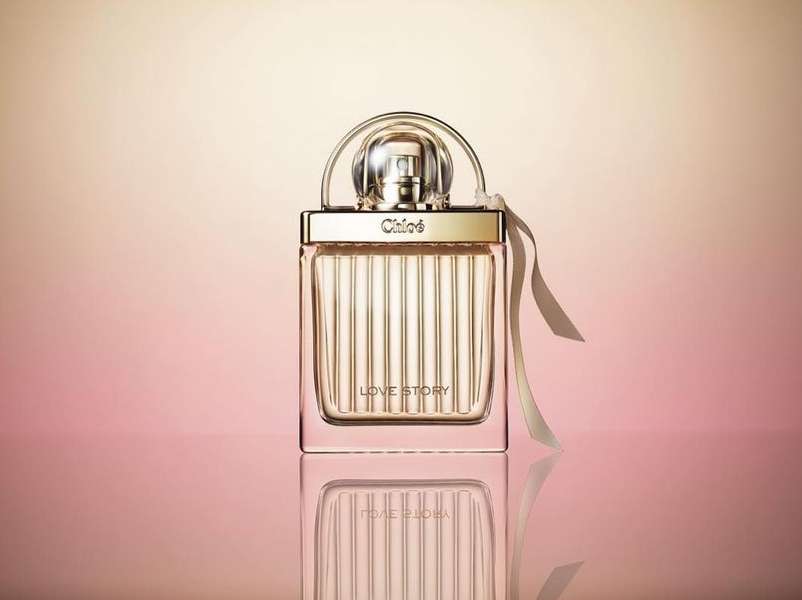 Chloé Love Story perfume bottle on a reflective surface with a pink and cream gradient background. Studio shot with clear lighting.