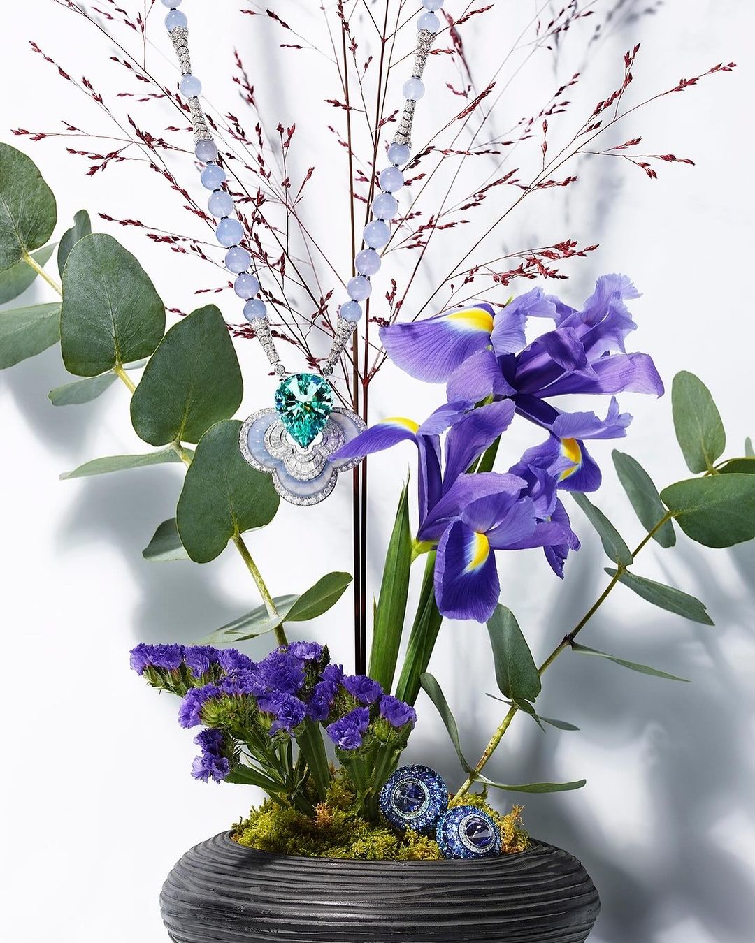 Elegant necklace with green and white pendant displayed among purple iris flowers and eucalyptus branches in a dark-grey textured vase.