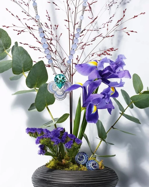 Elegant necklace with green and white pendant displayed among purple iris flowers and eucalyptus branches in a dark-grey textured vase.