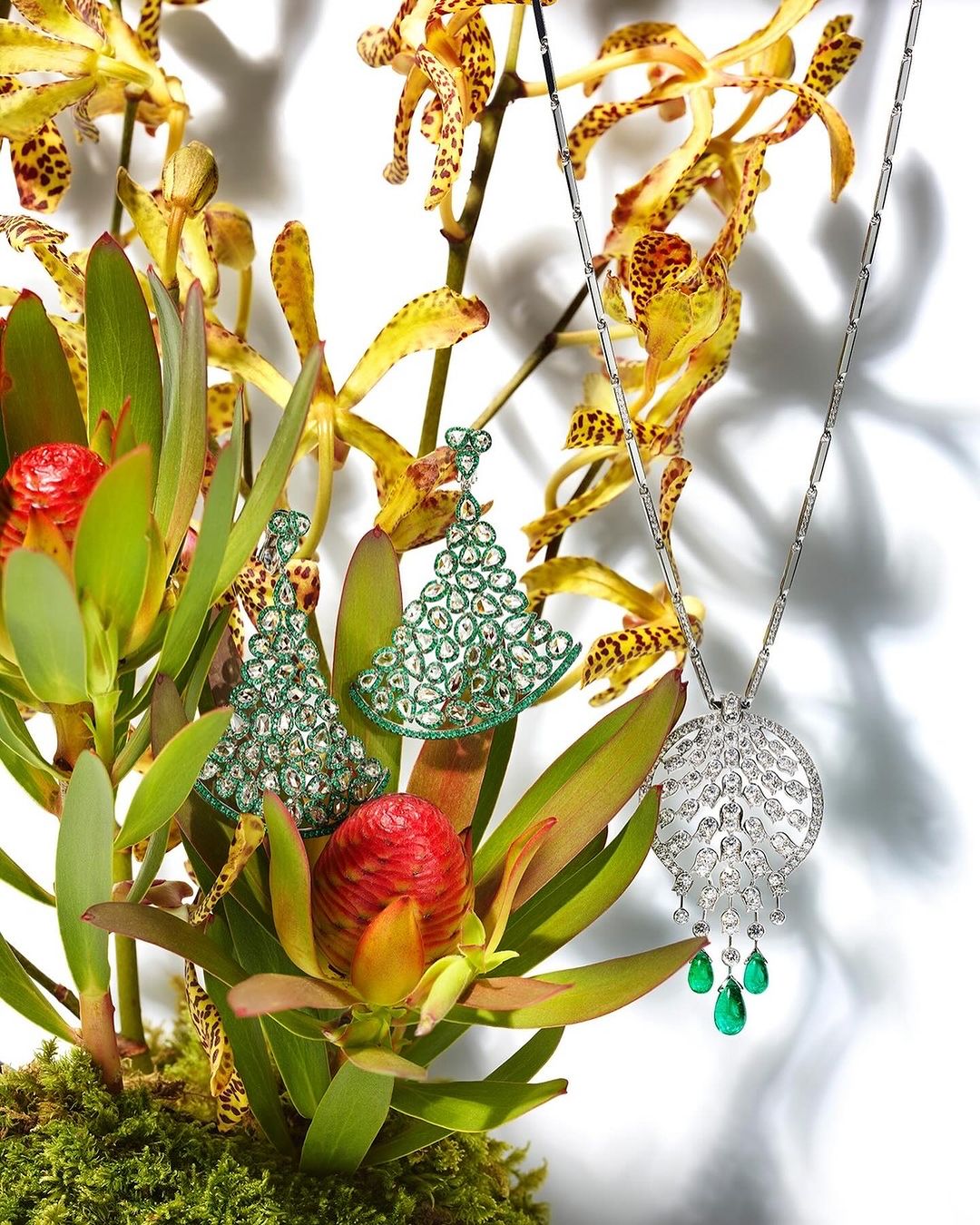 Elegant jewelry still life featuring diamond and emerald pendant necklace and green beaded earrings, set amidst green foliage and yellow orchids.