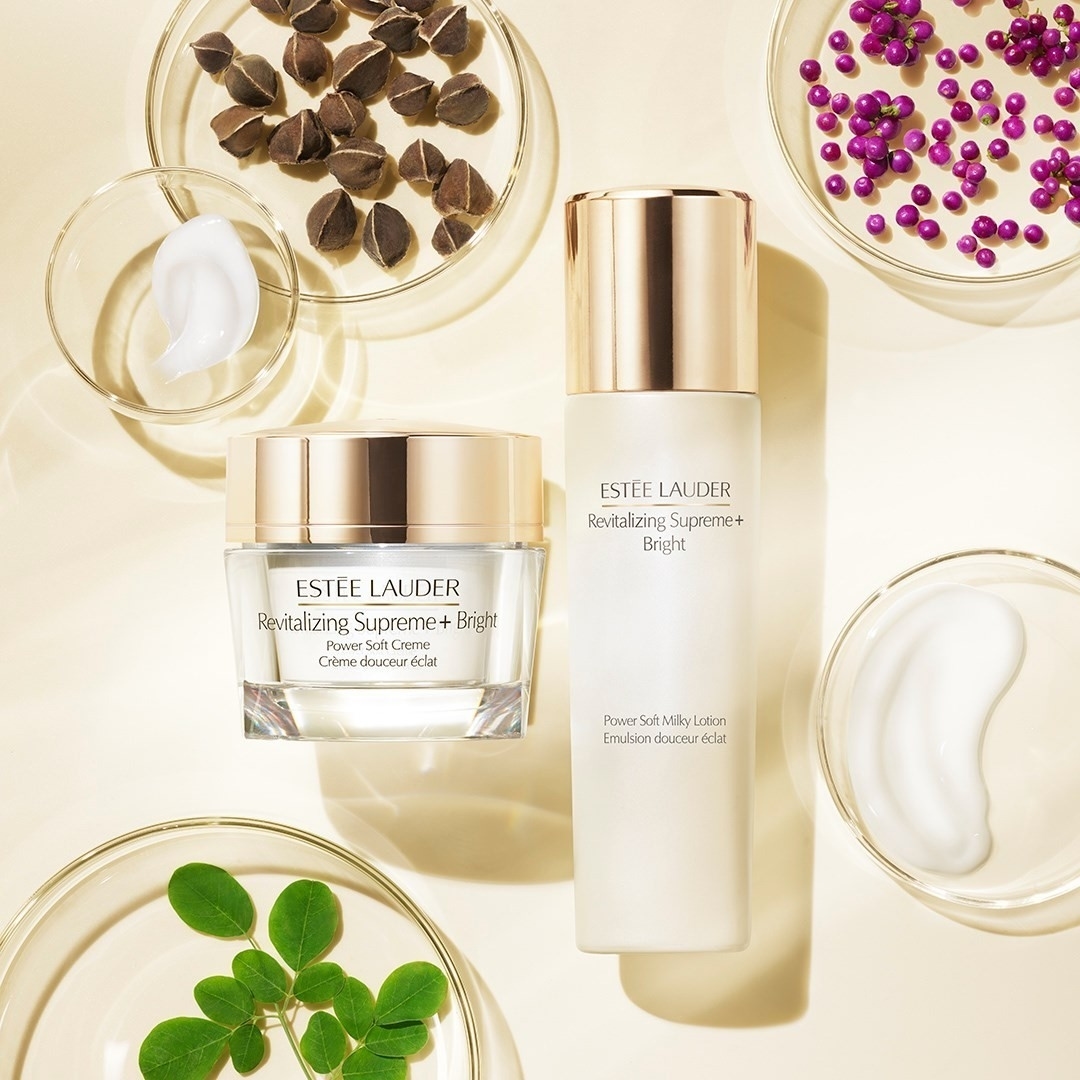 Two Estee Lauder Revitalizing Supreme+ Bright skincare products are displayed with petri dishes of botanical ingredients on beige.