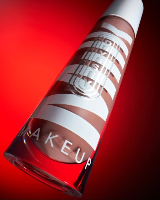 Product shot of a Milk Makeup product tube with white text on a clear container, filled with tan-colored makeup, set against a red background.