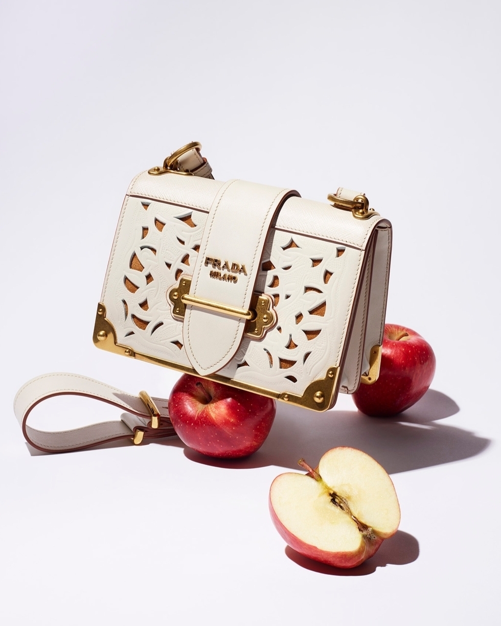 Prada handbag with cutout design, featuring a shoulder strap and gold hardware, resting on red apples against a plain white background.