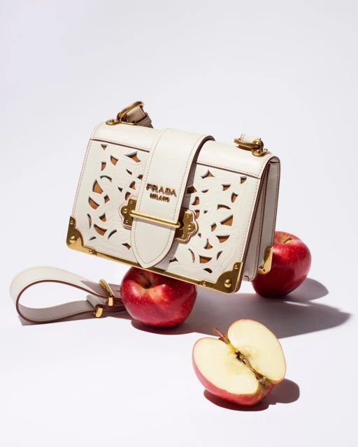 Prada handbag with cutout design, featuring a shoulder strap and gold hardware, resting on red apples against a plain white background.