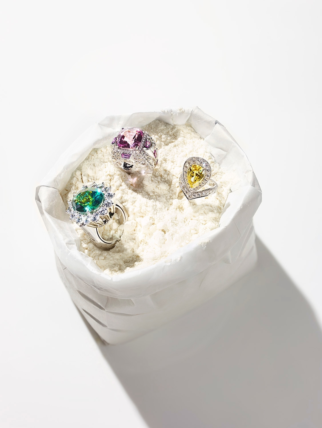 Three gemstone rings, with green, pink, and yellow gems, are displayed in a decorative white sack under bright studio lighting.