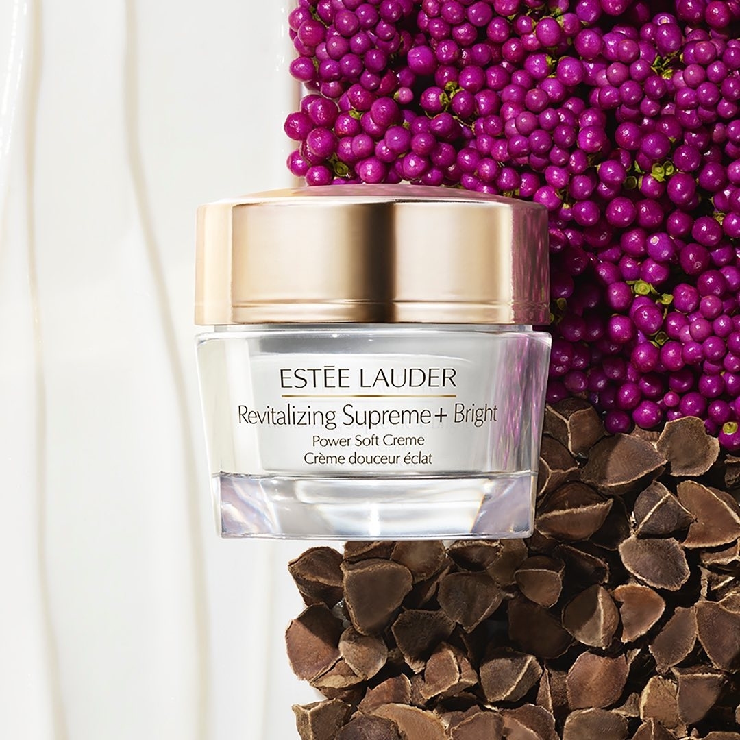 Close-up shot of Estee Lauder Revitalizing Supreme + Bright Power Soft Creme jar surrounded by pink berries and seeds on white surface.