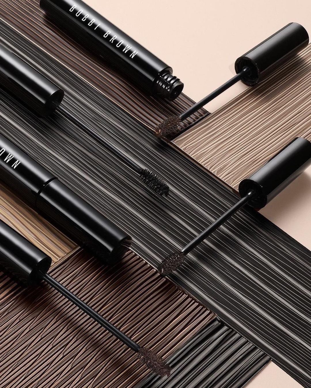 Close-up studio shot of multiple black Bobbi Brown mascara tubes and wands arranged on a textured brown and tan surface.