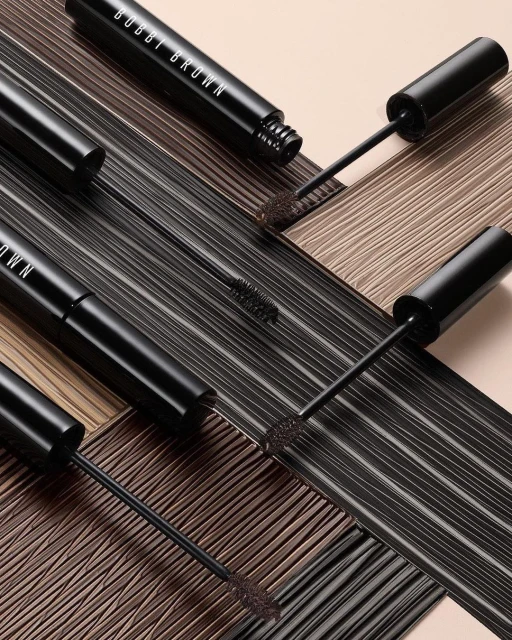 Close-up studio shot of multiple black Bobbi Brown mascara tubes and wands arranged on a textured brown and tan surface.