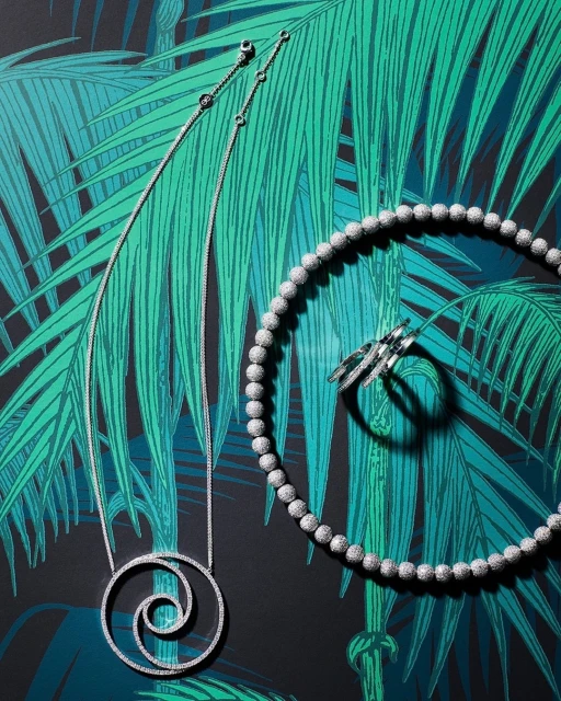 High-angle shot of a spiral pendant necklace, beaded necklace, and ring set against a green-and-black tropical leaf backdrop. Jewelry fashion.