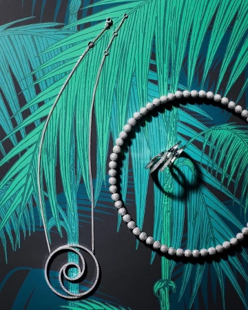 High-angle shot of a spiral pendant necklace, beaded necklace, and ring set against a green-and-black tropical leaf backdrop. Jewelry fashion.