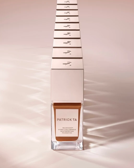 Patrick TA Major Skin Hydra-Luxe Luminous Skin Perfecting Foundation in clear glass packaging, studio shot on beige background.