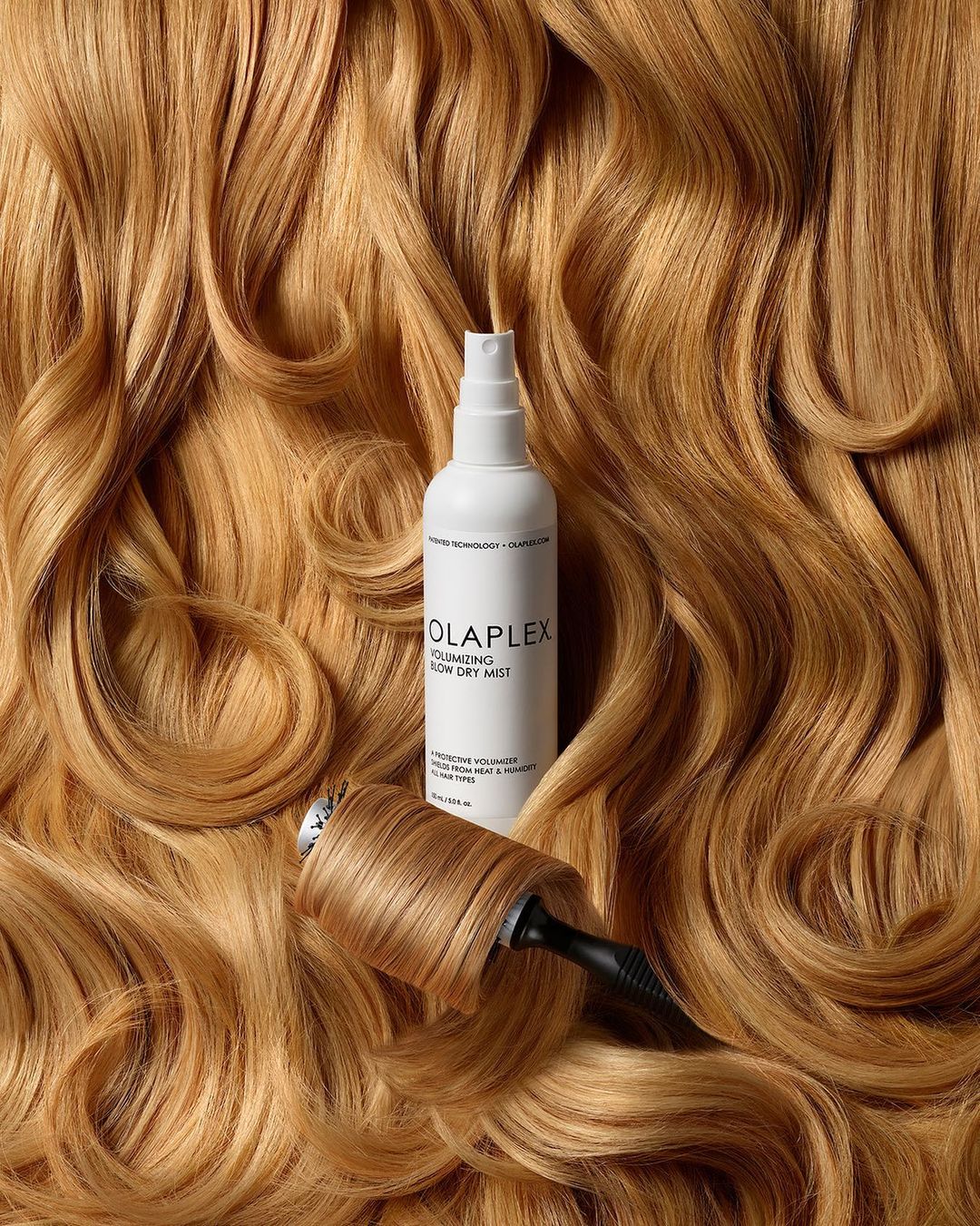 Product shot of Olaplex Volumizing Blow Dry Mist with tan-blonde wavy hair in background and wrapped around the base of the product.