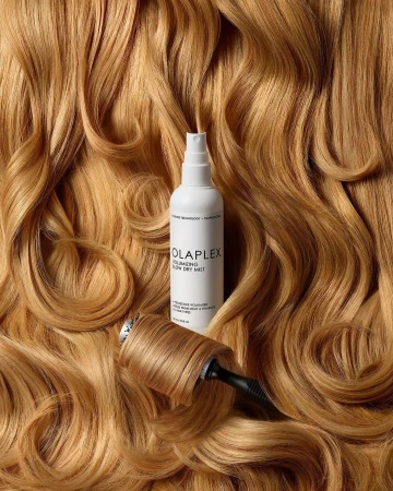 Product shot of Olaplex Volumizing Blow Dry Mist with tan-blonde wavy hair in background and wrapped around the base of the product.