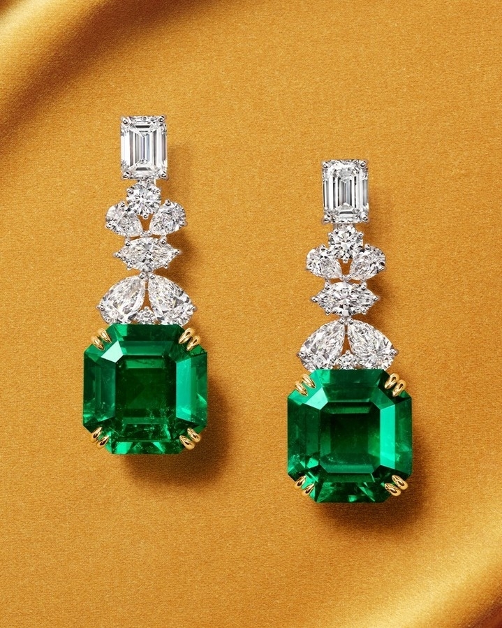 Pair of elegant emerald and diamond drop earrings on a gold background, showcasing luxury jewelry and detailed craftsmanship.