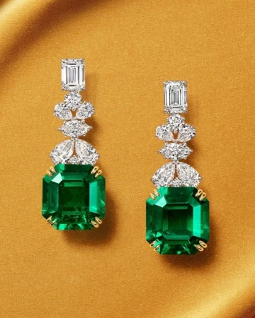 Pair of elegant emerald and diamond drop earrings on a gold background, showcasing luxury jewelry and detailed craftsmanship.