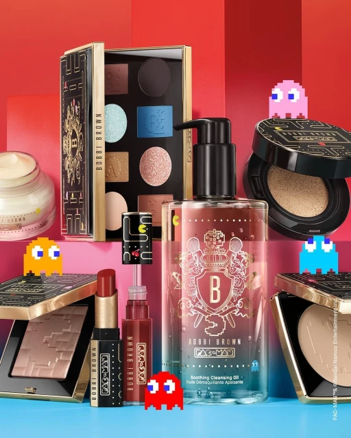 Bobbi Brown makeup collection featuring Pac-Man theme, including eyeshadow palette, cleansing oil, lipstick, and compacts on colorful background.