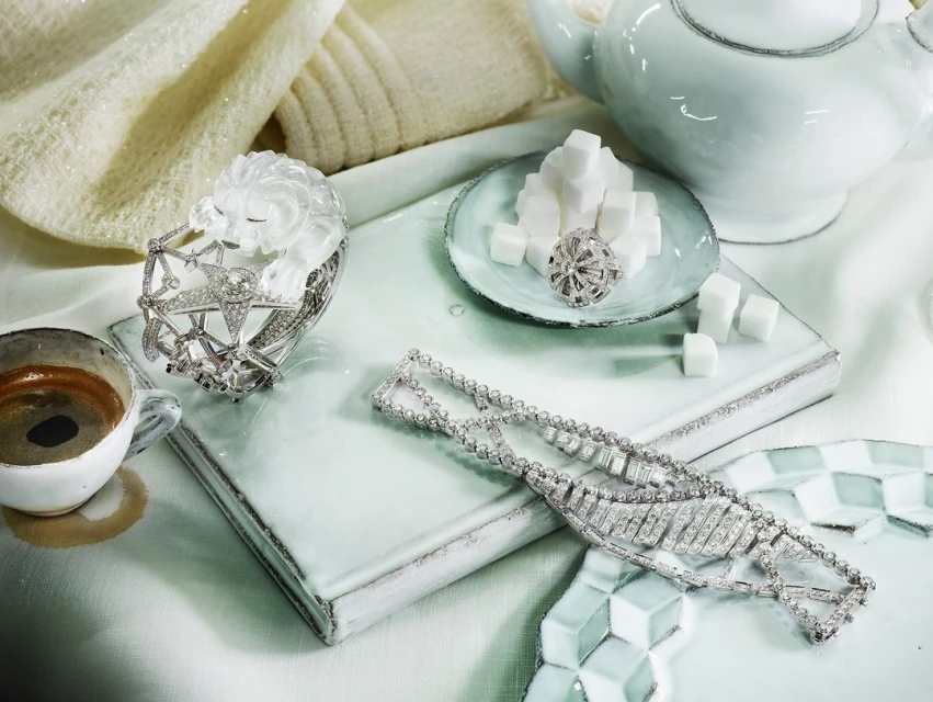 Still life of luxury jewelry on a tile surface with a cup of espresso, teapot, and sugar cubes in a light and bright setting.